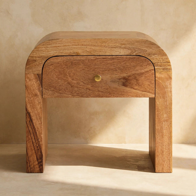 Darcy bedside table next to a bed, showing its rounded edges and mango wood grain