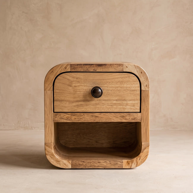 Small oak-ish bedside table with curved edges and one drawer beside a bed