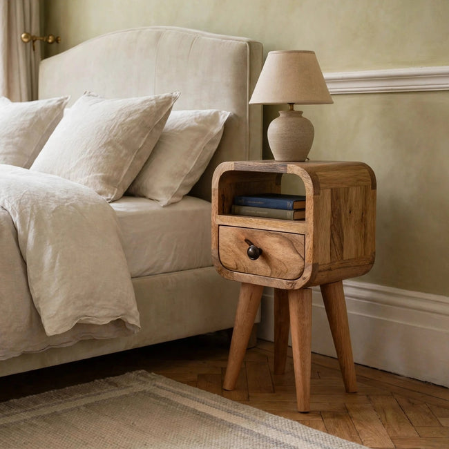 Mini oak-finish mango wood bedside table with curved edges, drawer and open slot, shown in a bedroom