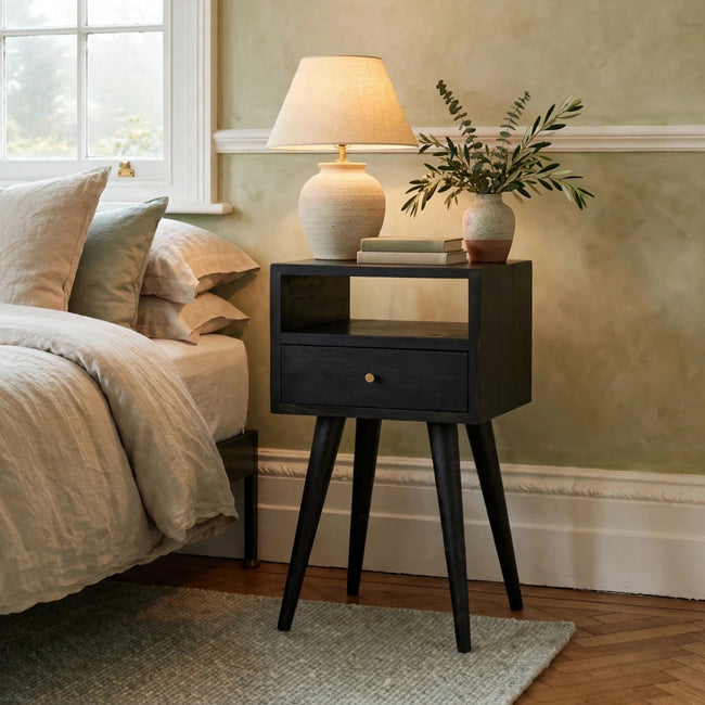 Mini ash black bedside table in solid mango wood standing next to a bed in a bedroom