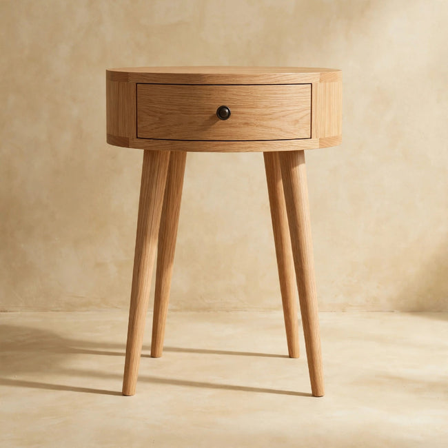 Round oak-ish bedside table with a drawer and shiny knob next to a bed